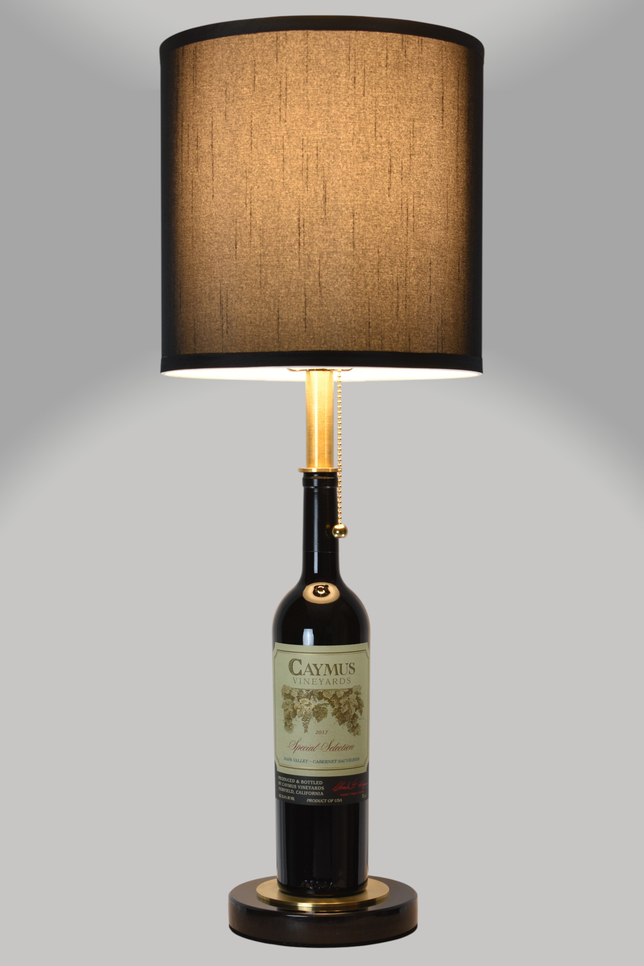 Wine bottle lamp made from a reclaimed 2017 Caymus Cabernet Sauvignon bottle, full view, light on, on a gray background