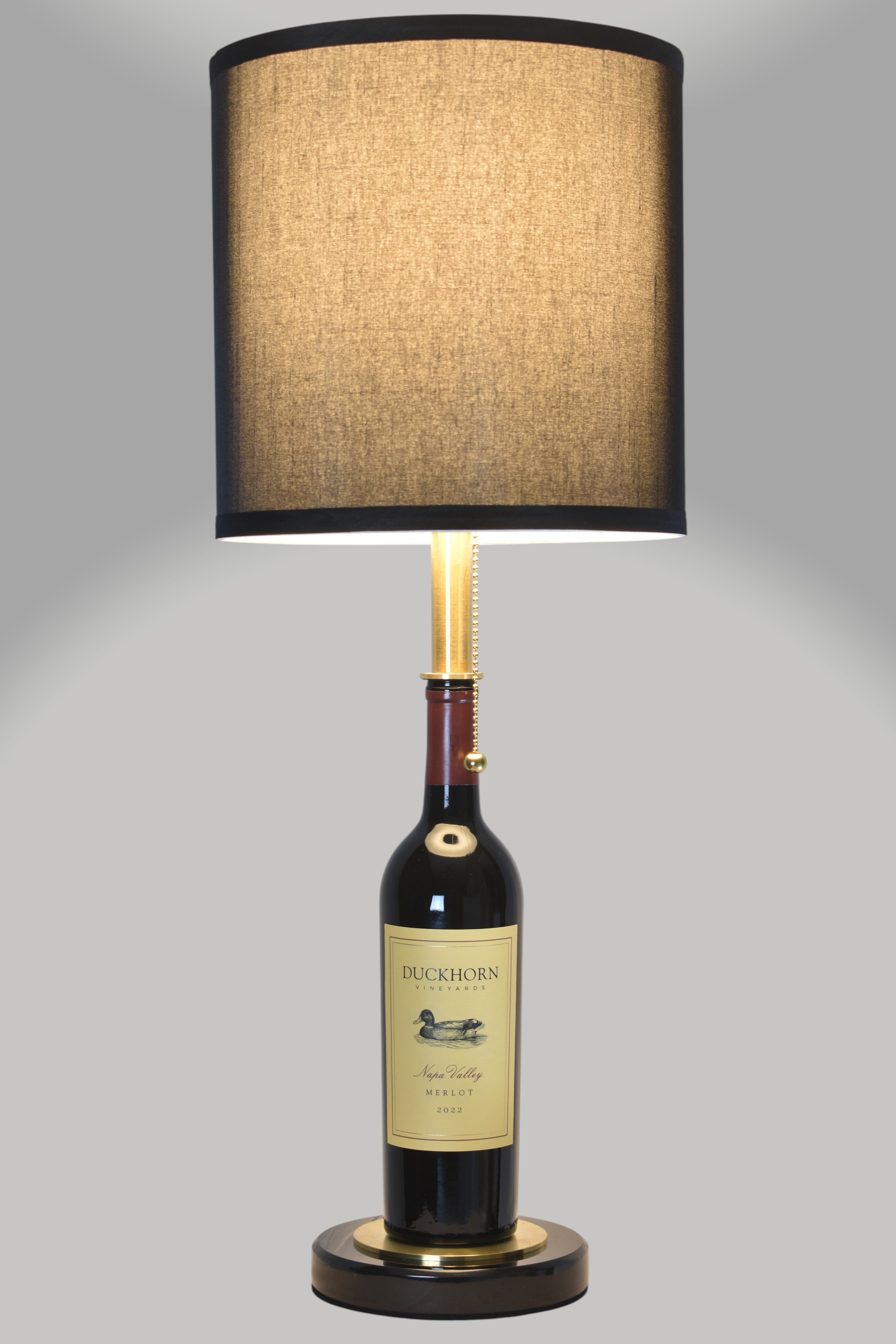 Wine bottle lamp made from a reclaimed 2022 Duckhorn Merlot bottle, full view, light on, on a gray background