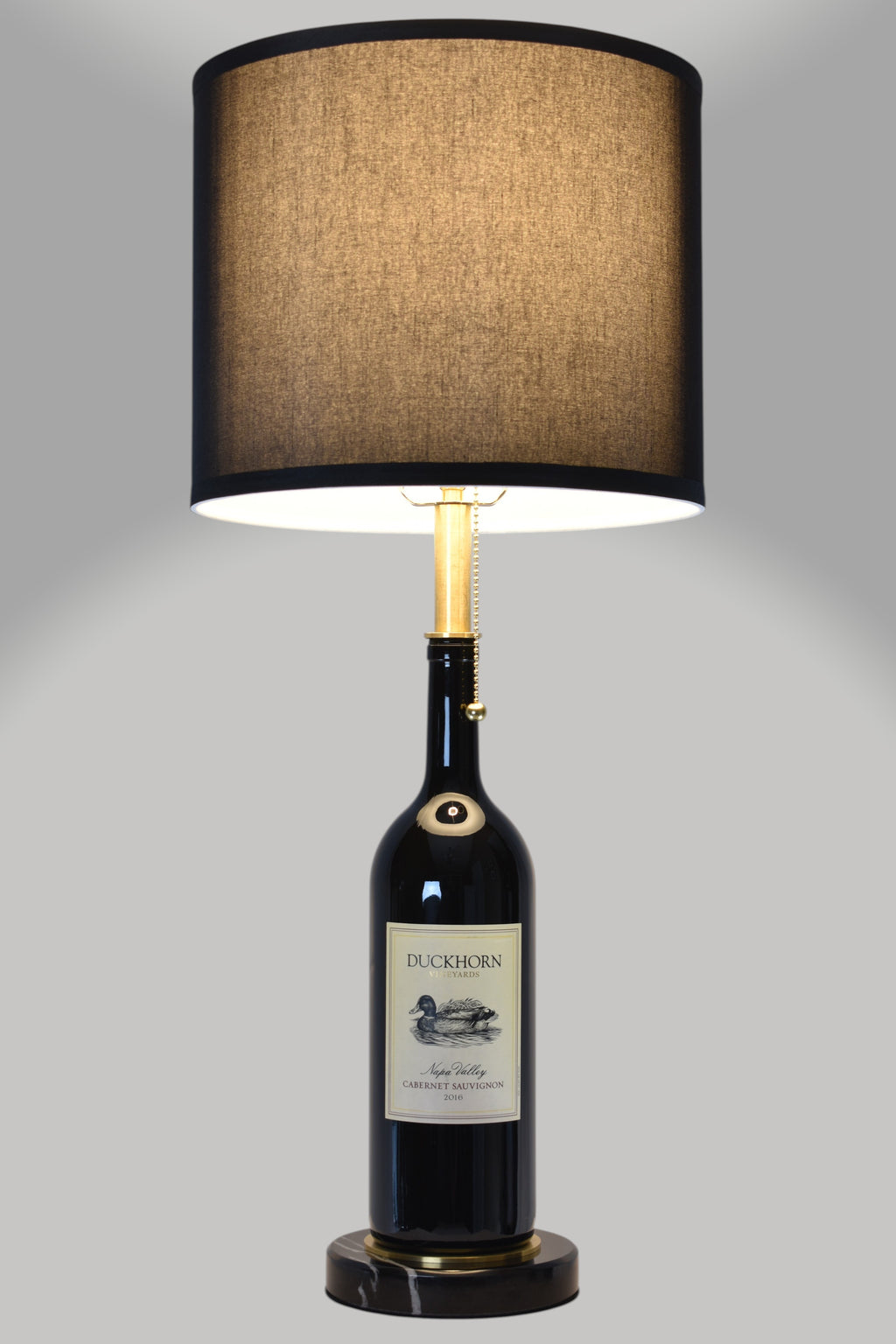 Wine bottle lamp made from a reclaimed 2016 Duckhorn Magnum Cabernet Sauvignon bottle, full view, light on, on a gray background