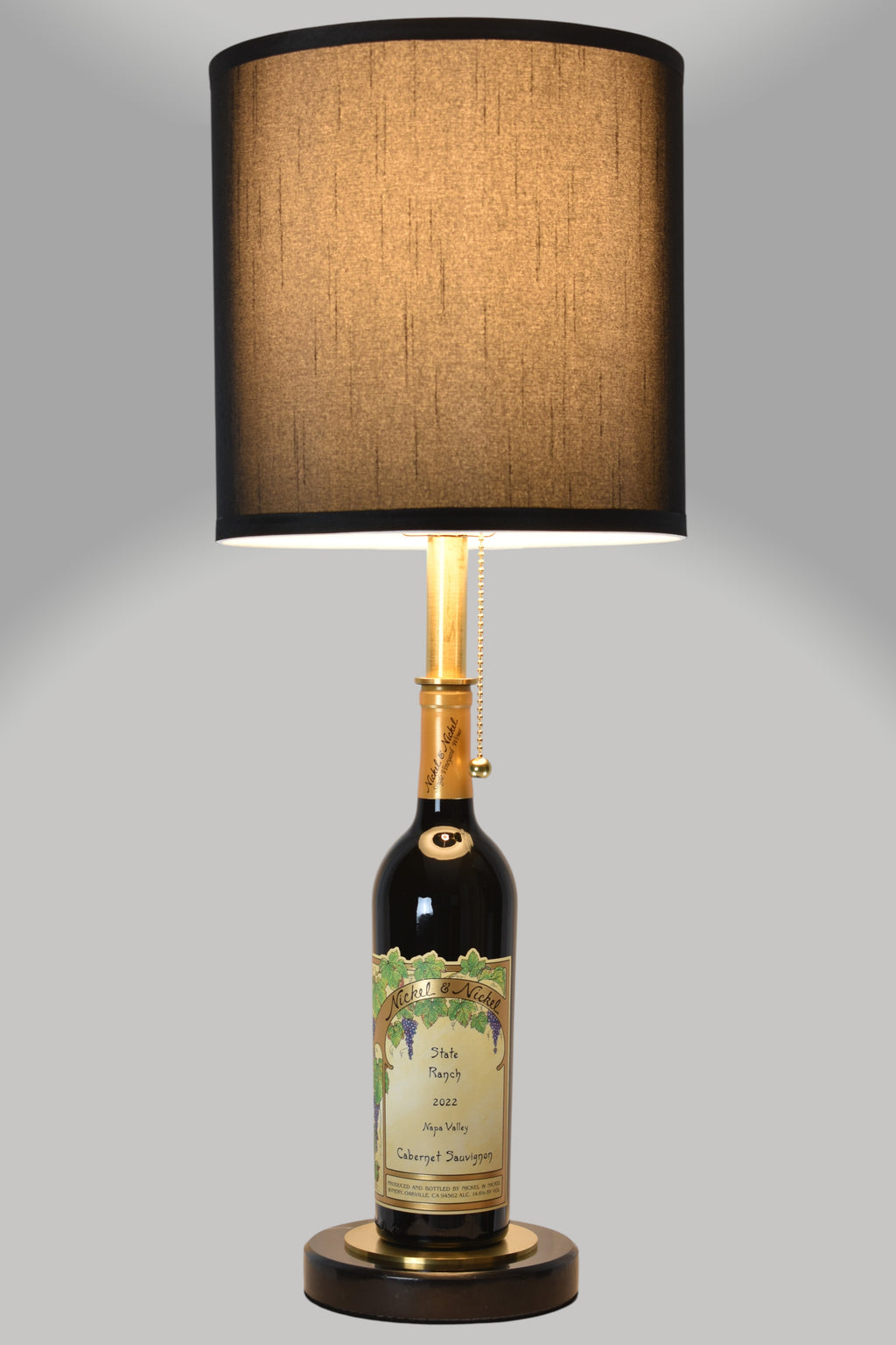 Wine bottle lamp made from a reclaimed 2022 Nickel & Nickel Cabernet Sauvignon bottle, full view, light on, on a gray background